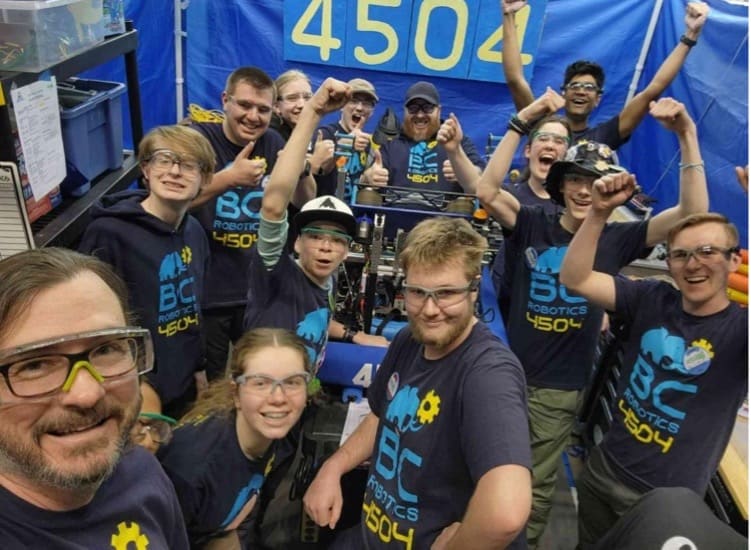 A group of enthusiastic robotics team members celebrating and posing for a photo with their robot, illustrating the supports of STEM education.