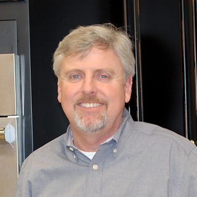 A man in a gray shirt standing in front of a black oven.